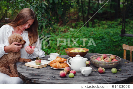 woman enjoying morning coffee in apple orchard with her pet poodle 91483008