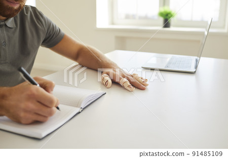 Student with splint braces on his broken fingers sitting at desk and writing in notebook Student with splint braces on his broken fingers sitting at desk and writing in notebook 91485109