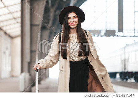Portrait of young beautiful woman traveler in casual coat and hat who is waiting for a train on the platform of the railway station. Portrait of young beautiful woman traveler in casual coat and hat who is waiting for a train on the platform of the railway station. 91487144
