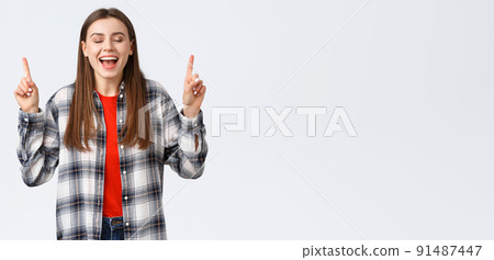 Lifestyle, different emotions, leisure activities concept. Dreamy and happy pretty young woman in checked shirt, dream came true, showing summer vacation resort, pointing fingers up with closed eyes 91487447