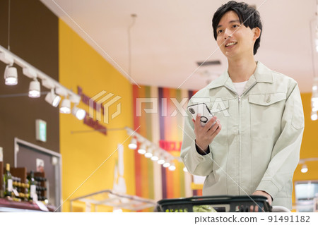一個穿著工作服的年輕人在超市購物,一隻手拿著智能手機 一個穿著工作服的年輕人在超市購物,一隻手拿著智能手機 91491182