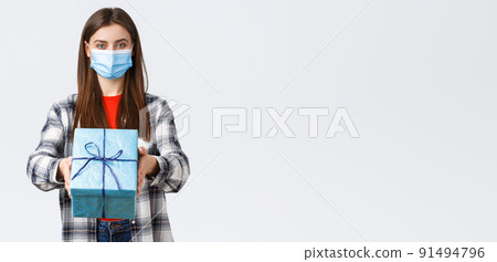 Covid-19, lifestyle, holidays and celebration concept. Cheerful young girl in medical mask stretch hand, handing birthday gift to you, smiling, congratulate with b-day, white background 91494796