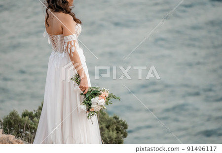 Gorgeous brunette bride posing with a bouquet on hotel terrace, looking at sea. Luxury destination, wedding makeup and hairstyle. Morning of the bride. Wedding concept 91496117
