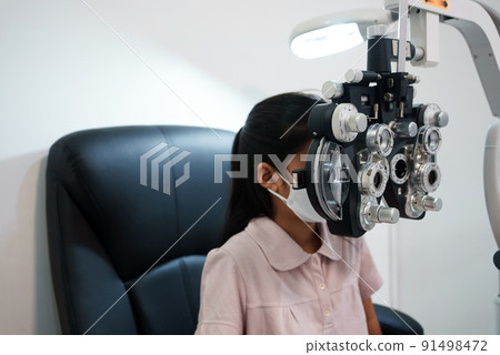 Ophthalmologist examining the eyes of an Asian girl in a clinic. They wear protective face masks. 91498472