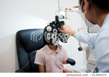 Ophthalmologist examining the eyes of an Asian girl in a clinic. They wear protective face masks. Ophthalmologist examining the eyes of an Asian girl in a clinic. They wear protective face masks. 91498474