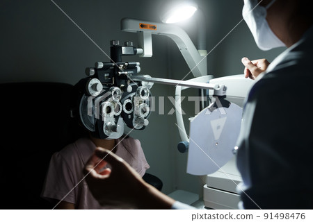 Ophthalmologist examining the eyes of an Asian girl in a clinic. They wear protective face masks. Ophthalmologist examining the eyes of an Asian girl in a clinic. They wear protective face masks. 91498476