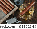 Top view of a Cuban cigar on a glass of whiskey or alcohol. Wooden table, lighter and cutter 91501343