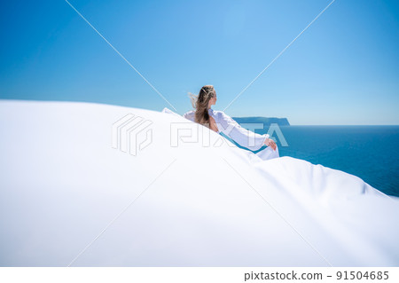 Blonde with long hair on a sunny seashore in a white flowing dress, rear view, silk fabric waving in the wind. Against the backdrop of the blue sky and mountains on the seashore. 91504685
