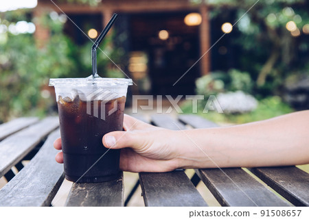 Cropped shot of someone hand touching a plastic cup of iced Americano (black coffee) on the wood plank table. Cropped shot of someone hand touching a plastic cup of iced Americano (black coffee) on the wood plank table. 91508657