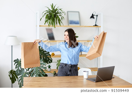 Attractive young woman stands in comfortable room and holding two paper bags on her hands. Beautiful trendy girl holds paper shopping bags and looks in camera with a smile Attractive young woman stands in comfortable room and holding two paper bags on her hands. Beautiful trendy girl holds paper shopping bags and looks in camera with a smile 91509449
