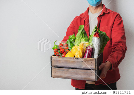 Young delivery man in protective face mask holding wooden box in hands. Man courier with shopping box. Safe online delivery from supermarket to home. COVID-19 coronavirus protection Young delivery man in protective face mask holding wooden box in hands. Man courier with shopping box. Safe online delivery from supermarket to home. COVID-19 coronavirus protection 91511731