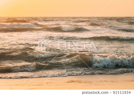 Sea ocean water surface with foaming small waves at sunset. Evening sunlight sunshine above sea. Ocean water foam splashes washing sandy beach. Crashing waves of sandy coastline. Natural sunset sky 91511934