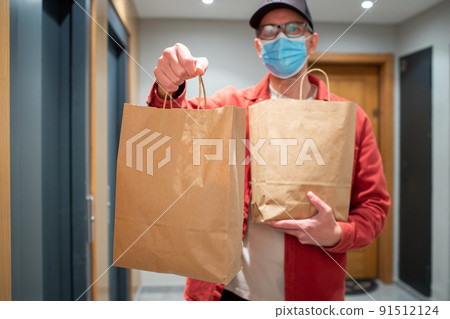 Delivery man in protective mask holding paper bag with food in the entrance. The courier gives the box with fresh vegetables and fruits to the customer 91512124