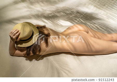 Naked beautiful woman nudist in straw hat lying and sunbathes in a shadow under palm tree branches. Female relaxation on the sand beach at summer vacation. Top view 91512243