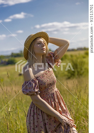 Happy beautiful girl, holding her hand on her hat, admires the beauty in the meadow at sunset. 91513701