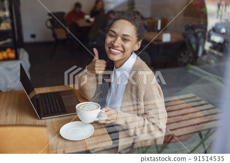 Beautiful stylish African American woman showing thumb up while working on laptop in a coffee shop 91514535