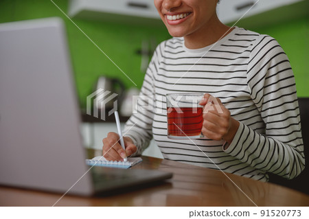 Close up shot of happy young woman holding cup of tea and writing in notepad using laptop 91520773