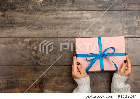Cropped view of woman holding wrapped christmas gift with blue bow. Female hands holding gift box on wooden rustic background 91528244