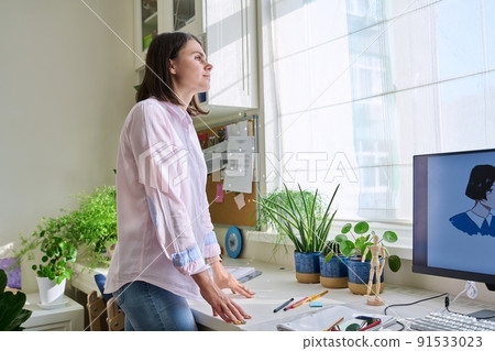 Young woman looking out the window standing near workplace in home office 91533023
