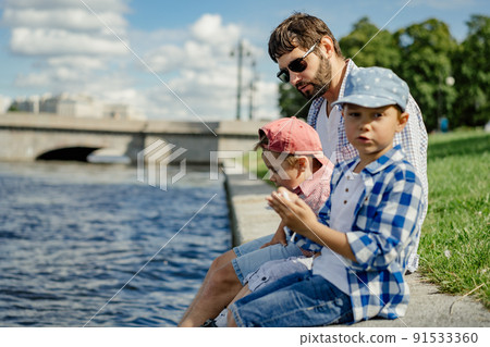 father and two boys sitting on the bank of Neva river spending time together, Saint-petersburg, Russia. 91533360