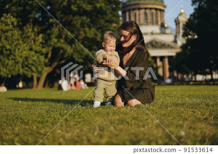 Caucasian mother of little son sitting on grass in front of st isaac cathedral helping boy stand holding by hand. Caucasian mother of little son sitting on grass in front of st isaac cathedral helping boy stand holding by hand. 91533614