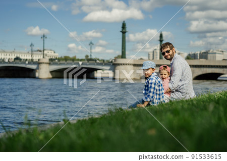 father and two boys sitting on the bank of Neva river spending time together, Saint-petersburg, Russia. 91533615