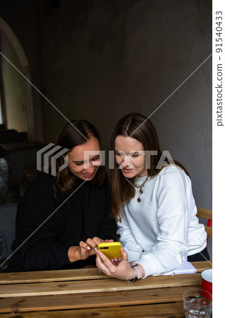 Two young women communicate and look into smartphone in cafe on terrace Two young women communicate and look into smartphone in cafe on terrace 91540433