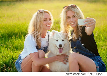two beautiful and charming blonde twins woman with a white fluffy samoyed dog sitting and making selfie on the phone in the park 91543559