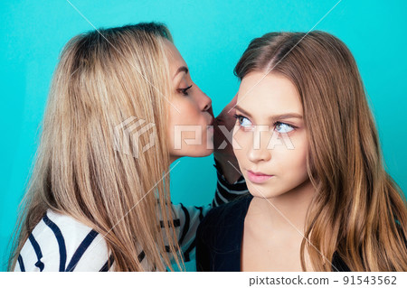 portrait of two young attractive happy women with blonde hair and makeup eavesdrops whispers a secret mystery in the studio on a blue background. the concept of gossip and confidentiality portrait of two young attractive happy women with blonde hair and makeup eavesdrops whispers a secret mystery in the studio on a blue background. the concept of gossip and confidentiality 91543562