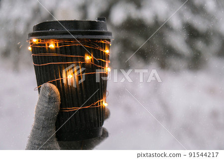 Female hand in winter warm gloves holding black eco paper cup with garland shiny light. Creative trendy zero waste recycle cup. Mockup. Hot warming drink to go in winter day. Take away concept 91544217