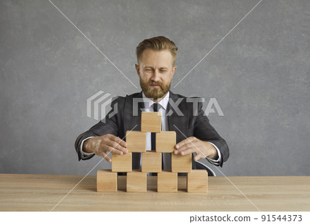 Business leader sitting at office desk and building pyramid out of wooden blocks Business leader sitting at office desk and building pyramid out of wooden blocks 91544373