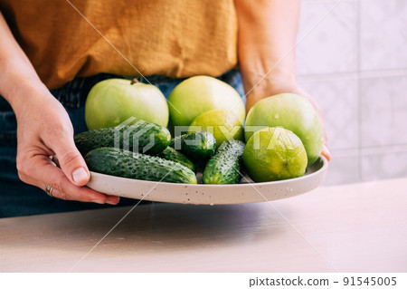 A woman holds in her hands fresh green vegetables fruits in large plate, light background copy space A woman holds in her hands fresh green vegetables fruits in large plate, light background copy space 91545005