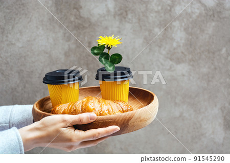 Two yellow paper cups for coffee or tea and Fresh croissant on a wooden tray. Takeaway order in women's hands. Dandelion flower in the lid of a container for a romantic breakfast. Copy space for text 91545290