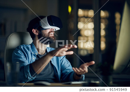 Excited young bearded man sitting in front of computer and gesturing hands while testing new app via VR device 91545949