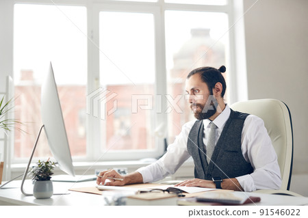 Concentrated young bearded businessman with ponytail sitting at office table and using computer while analyzing online sales 91546022