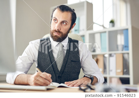 Serious thoughtful young bearded lawyer in gray waistcoat and white shirt sitting at table and making notes in planner while looking at computer monitor 91546023