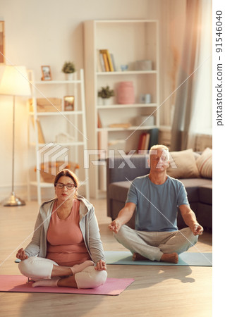 Serene senior couple with crossed legs sitting on yoga mats in living room and meditating with closed eyes 91546040
