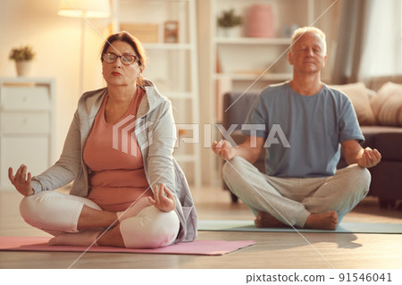 Serene relaxed senior couple with closed eyes sitting with crossed legs on floor in living room and practicing meditation Serene relaxed senior couple with closed eyes sitting with crossed legs on floor in living room and practicing meditation 91546041