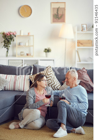 Senior brown-haired woman in glasses sitting on carpet in living room and chatting with husband while drinking wine Senior brown-haired woman in glasses sitting on carpet in living room and chatting with husband while drinking wine 91546435