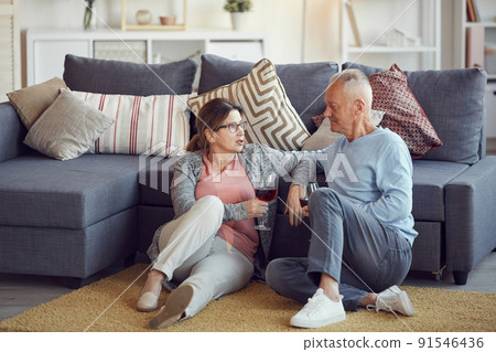 Content relaxed senior couple in casual clothing sitting on floor in living room and drinking red wine while talking Content relaxed senior couple in casual clothing sitting on floor in living room and drinking red wine while talking 91546436