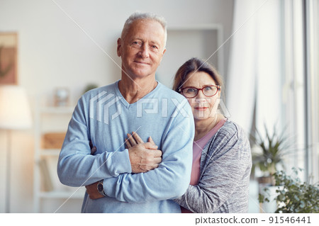 Smiling beautiful senior couple standing in living room and hugging while posing for portrait Smiling beautiful senior couple standing in living room and hugging while posing for portrait 91546441