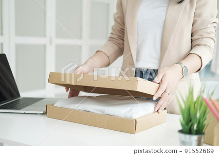 Asian female clothes shop owner folding a t-shirt and packing in a cardboard parcel box. Asian female clothes shop owner folding a t-shirt and packing in a cardboard parcel box. 91552689