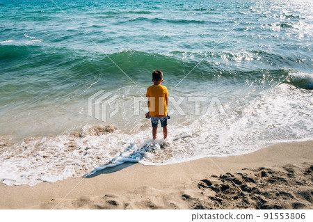 a boy in yellow clothes stands on the sea beach in front of a rolling wave in the sunset rays of the sun a boy in yellow clothes stands on the sea beach in front of a rolling wave in the sunset rays of the sun 91553806