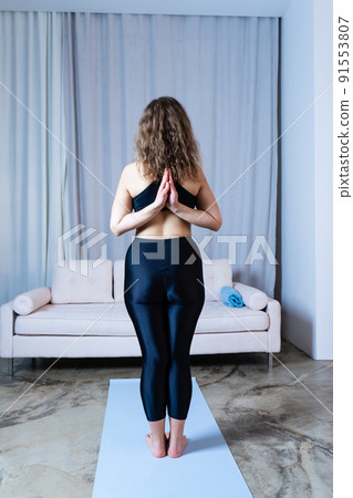 Young healthy beautiful woman in sports top and leggings practicing yoga at home, standing in pose on yoga mat, namaste 91553807