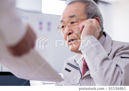 一位忙碌的老人一邊用商務智能手機交談一邊閱讀文件 一位忙碌的老人一邊用商務智能手機交談一邊閱讀文件 91556861