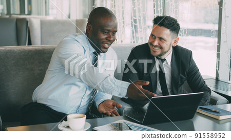 Cheerful African American businessman in formal clothes discussing business project with his caucasian colleague on his laptop in stylish cafe during lunch break 91558952
