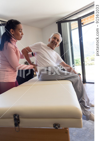Biracial female physiotherapist giving back massage therapy to caucasian senior man at home 91561033