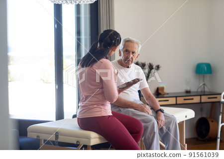 Biracial female physiotherapist with digital tablet discussing with caucasian senior man 91561039