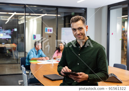 Portrait of confident young caucasian businessman with tablet pc while colleagues in background 91561322