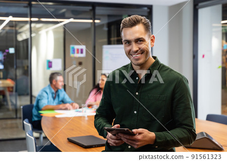 Portrait of smiling young caucasian businessman with tablet pc while colleagues in background Portrait of smiling young caucasian businessman with tablet pc while colleagues in background 91561323